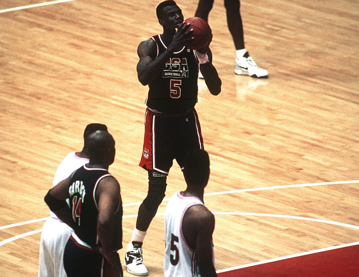 A U.S. men’s basketball player in a dark USA jersey shoots a free throw during a 1992 Olympic game in Barcelona while teammates and opponents line up along the lane.